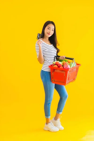 黄色の隔離された背景にデパートのスーパーマーケットからのクレジットカードでバスケットの食料品やカートを持つ肖像画の美しい若いアジアの女性 — ストック写真