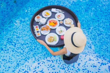 Asyalı güzel bir kadının portresi yüzme havuzunda yüzen kahvaltı tepsisiyle mutlu bir gülümseme