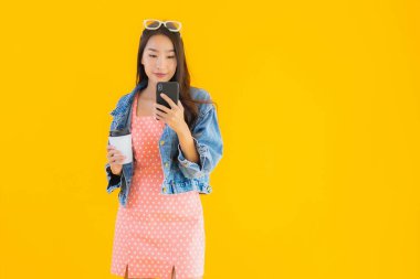 Kahve fincanı ve sarı izole edilmiş arka planda akıllı cep telefonlu güzel Asyalı kadın portresi.