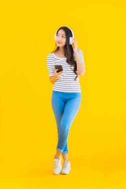 Güzel bir Asyalı kadın portresi, sarı izole edilmiş arka planda müzik dinlemek için kulaklıklı akıllı cep telefonu kullanır.