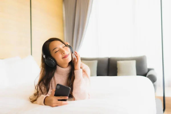 Portrait beautiful young asian woman using mobile phone with head phone for listen music in bedroom interior