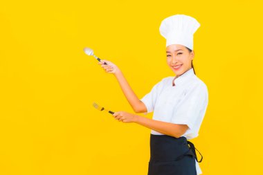 Güzel bir portre genç Asyalı kadın aşçı ya da aşçı üniforması giyiyor sarı izole arka planda çatal ve kaşıkla.