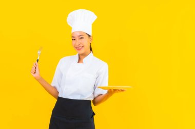 Güzel bir portre genç Asyalı kadın aşçı ya da aşçı üniforması giyiyor sarı izole arka planda çatal ve kaşıkla.