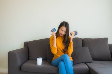 Genç Asyalı kadının oturma odasında online alışveriş yapmak için akıllı cep telefonu ve kredi kartı olan dizüstü bilgisayarı kullandığı portre.