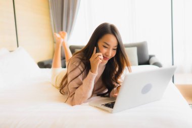 Yatak odasının iç kısmında dizüstü bilgisayarı ve cep telefonu olan güzel Asyalı kadın portresi.