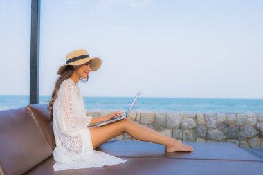 Plaj denizi çevresinde çalışmak için dizüstü bilgisayarı kullanan Asyalı genç bir kadının portresi.