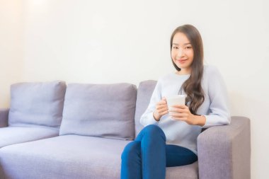 Oturma odasındaki kanepede kahve fincanı taşıyan güzel Asyalı kadın portresi.