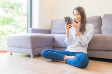 Güzel bir Asyalı kadın oturma odasındaki koltukta akıllı cep telefonu kullanıyor.