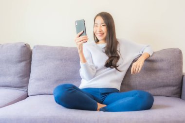 Güzel bir Asyalı kadın oturma odasındaki koltukta akıllı cep telefonu kullanıyor.