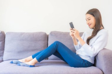 Güzel bir Asyalı kadın oturma odasındaki koltukta akıllı cep telefonu kullanıyor.