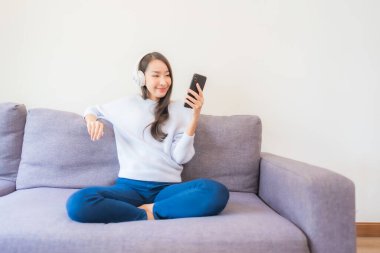 Güzel bir Asyalı kadın oturma odasında müzik dinlemek için akıllı cep telefonu kulaklığı kullanıyor.