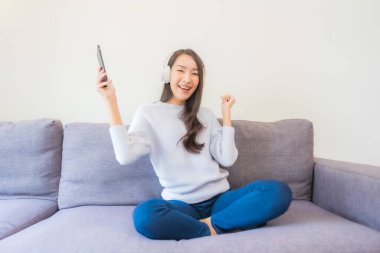 Güzel bir Asyalı kadın oturma odasında müzik dinlemek için akıllı cep telefonu kulaklığı kullanıyor.