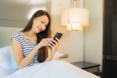 Güzel bir Asyalı kadın yatak odasının içinde akıllı cep telefonu kullanıyor.