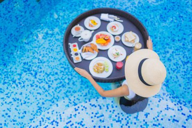 Asyalı güzel bir kadının portresi yüzme havuzunda yüzen kahvaltı tepsisiyle mutlu bir gülümseme