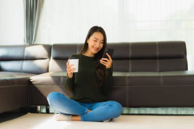 Güzel bir Asyalı kadın portresi oturma odasının iç tarafında kanepeli bir kahve fincanı ve akıllı cep telefonu kullanıyor.