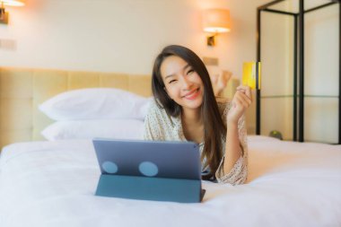 Güzel bir Asyalı kadının portresi yatak odasının içinde kredi kartıyla tablet kullanıyor.
