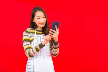 Güzel bir Asyalı kadın portresi kırmızı izole edilmiş arka planda akıllı cep telefonuyla önlük giyiyor.