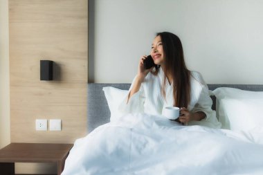 Güzel Asyalı genç kadınların portresi. Mutlu bir gülümseme. Yatak odasındaki yatakta kahve ve cep telefonuyla.