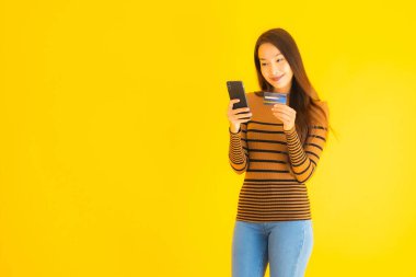 Asyalı güzel bir kadının portresi, sarı izole edilmiş arka planda online alışveriş için akıllı cep telefonu ya da kredi kartı kullanıyor.
