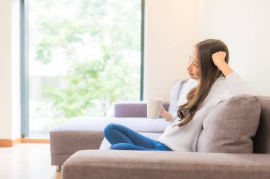 Oturma odasındaki kanepede kahve fincanı taşıyan güzel Asyalı kadın portresi.