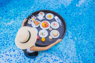 Asyalı güzel bir kadının portresi yüzme havuzunda yüzen kahvaltı tepsisiyle mutlu bir gülümseme