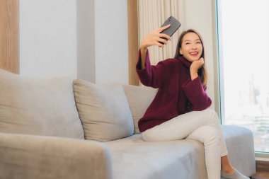 Güzel bir Asyalı kadın oturma odasındaki koltukta akıllı cep telefonu kullanıyor.