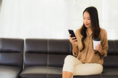 Güzel bir Asyalı kadın portresi. Kanepenin iç tarafında cep telefonu ya da kahve fincanı ile cep telefonu kullanıyor.