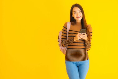 Çantalı güzel Asyalı kadın sarı izole edilmiş arka planda akıllı cep telefonu ya da cep telefonu kullanıyor.