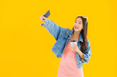 Kahve fincanı ve sarı izole edilmiş arka planda akıllı cep telefonlu güzel Asyalı kadın portresi.