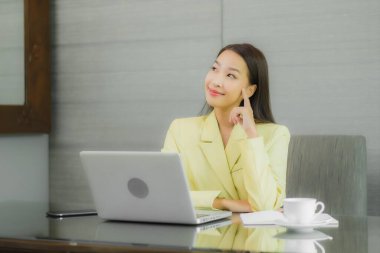 Asyalı güzel bir kadının portresi iç odasındaki çalışma masasında çalışan akıllı cep telefonlu bir bilgisayar dizüstü bilgisayar kullanıyor.