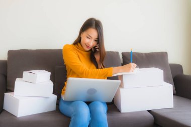 Asyalı genç bir kadının portresi, oturma odasındaki koltukta paket kutusuyla çalışmak için dizüstü bilgisayar kullanıyor.