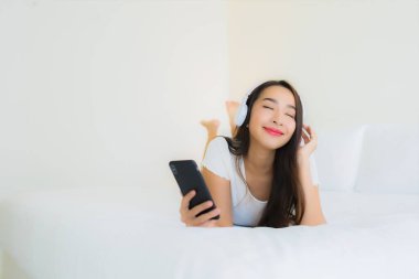 Asyalı güzel bir kadının portresi mutlu bir gülümseme akıllı cep telefonu ve kulaklıkla yatak odasının içinde müzik dinlemek için.