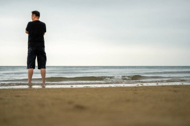 Uzun boylu Asyalı bir adam sahilde dikiliyor.