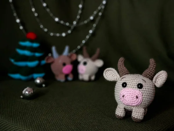 Beige crocheted toy bull cow in the foreground. In the background a green background, two other toys, Christmas tree and festive decorations. — Stock Photo, Image