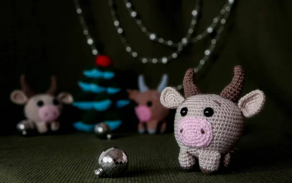 Beige crocheted toy bull cow in the foreground. In the background a green background, two other toys, Christmas tree and festive decorations. — Stock Photo, Image