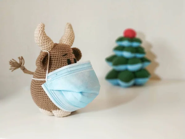 Crochet soft toy bull cow in blue medical protective mask. On a light background with a toy fir tree. New Year Christmas 2021 — Stock Photo, Image