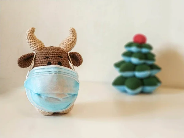 Crochet soft toy bull cow in blue medical protective mask. On a light background with a toy fir tree. New Year Christmas 2021 — Stock Photo, Image