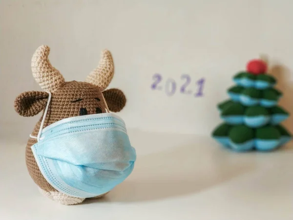 Crochet soft toy bull cow in blue medical protective mask. On a light background with a toy fir tree, blurry inscription 2021. New Year Christmas 2021 — Stock Photo, Image