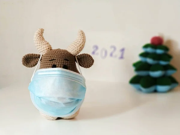 Crochet soft toy bull cow in blue medical protective mask. On a light background with a toy fir tree, blurry inscription 2021. New Year Christmas 2021 — Stock Photo, Image