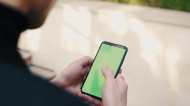 Açık havada yeşil ekranlı bir telefon kullanan bir adamın omuz üstünden. Yakışıklı adam şehrin dışındaki akıllı telefon uygulamalarında kayıyor..