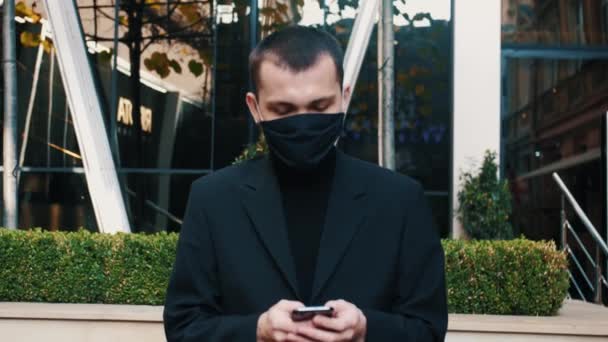 Joven hombre de negocios caucásico en máscara médica caminando por la calle y mensajes de texto en el teléfono móvil. Peatón masculino en protección respiratoria paseando al aire libre y tocando o desplazándose en el teléfono inteligente. — Vídeos de Stock