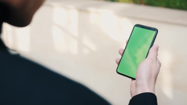 Sobre o ombro tiro de homem usando um telefone com uma tela verde ao ar livre. Homem bonito rolando através de aplicativos de smartphones fora da cidade. — Vídeo de Stock