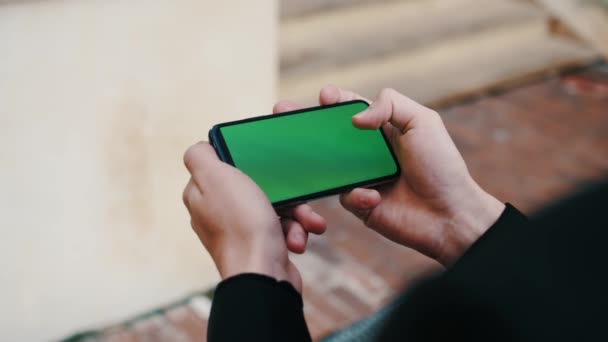 Человек, использующий смартфон в горизонтальном режиме с зеленым макетом экрана за пределами городского делового офиса — стоковое видео
