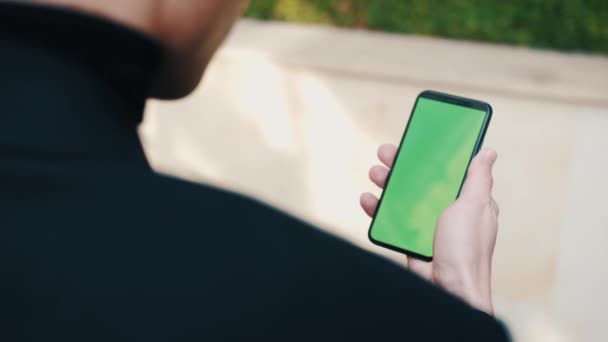 Over de schouder van de mens met behulp van een telefoon met een groen scherm buiten. Knappe man scrollen door smartphone apps buiten in de stad. — Stockvideo