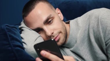 Happy Young Kafkasyalı adam evinde bir koltukta otururken akıllı telefon kullanıyor. Man İnternet 'e Bakar, Video İzler ve Evde Sosyal Ağ Kullanır.