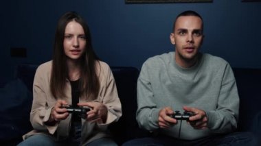 Heyecanlı çift evdeki TV karşısında kanepede otururken video oyunu oynuyor. Genç adam ve kadın boş zamanlarında joystickleri tutuyorlar..