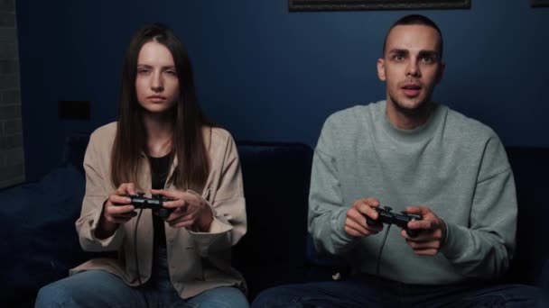 Nadšený pár hraje videohry, zatímco sedí na pohovce před televizí doma. Mladý muž a žena drží joysticks zatímco tráví volný čas. — Stock video