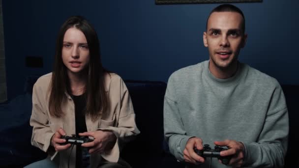 Emocionada pareja jugando videojuegos mientras está sentada en el sofá frente a la televisión en casa. Joven hombre y mujer sosteniendo joysticks mientras pasa tiempo libre. — Vídeo de stock