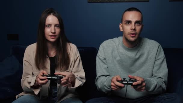 Emocionada pareja jugando videojuegos mientras está sentada en el sofá frente a la televisión en casa. Joven hombre y mujer sosteniendo joysticks mientras pasa tiempo libre. — Vídeos de Stock