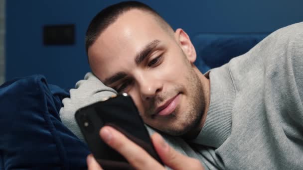 Щасливий молодий чоловік використовує смартфон, сидячи на дивані вдома. Чоловік переглядає Інтернет, дивиться відео та використовує соціальні мережі вдома . — стокове відео